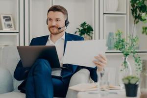 Horizontal shot of serious bearded male employee uses modern laptop computer for distance working wears headphones with microphone does paper work prepares for virtual conference with partner photo