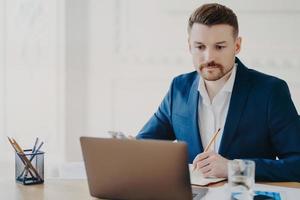 Serious bearded man marketer involved in working process makes notes with pencil looks attentively at laptop computer wears blue formal suit poses at coworking space writes organisation plan photo