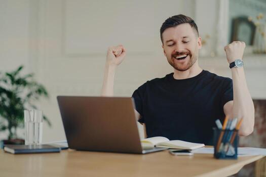 Excited cheerful bearded man with joyful expression clenches fists celebrates success dressed casually poses at desktop in front of opened laptop satisfied with results of work. Yes great job photo