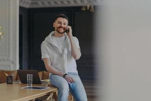 Happy bearded male freelancer works in home office room dressed in casual t shirt and jeans has telephone conversation discusses working issues has cheerful expression works remotely from home photo