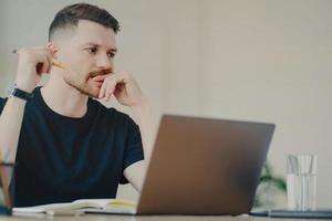 Serious bearded man student enjoys studying process has thoughtful expression works freelance dressed in casual black t shirt writes down notes uses modern laptop computer poses in coworking space photo