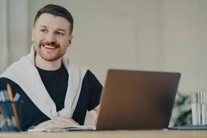 Happy bearded man office worker writes organisation plan works remotely makes notes of info sits in front of opened laptop looks away dressed in casual t shirt and jumper tied over shoulders photo