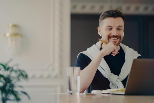 Horizontal shot of happy busy male student prepares project work focused at lpatop computer uses wireless earphones creats interesting content on own website writes down information with pencil photo