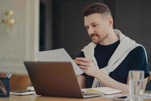 Concentrated man reads information or data from paper documents works on freelance poses at desktop against cozy interior dressed in casual clothes busy working. Male entrepreneur at coworking space photo