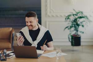 Happy bearded young man studies indoor with modern gadgets writes down notes from internet website makes records in notebook holds smartphone poses at coworking space cozy interior. Hipster student photo