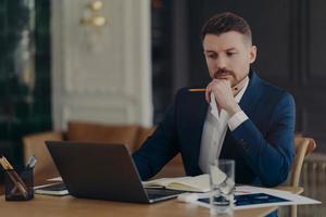 Serious male entrepreneur holds chin makes notes in diary works on laptop computer makes money online dressed formally writes information. Confident managing director involved in working process photo