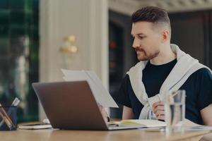 Smart handsome man checks information on papers works remotely sits at home office uses modern laptop computer writes down in notebook dressed in casual clothes. Freelance work and technology photo