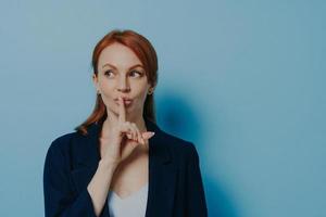 Young mysterious redhead europian female asking to keep silence and looking aside, isolated on blue photo