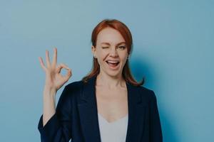 Young positive redhead woman in classic outfit showing okay gesture with hand and winking photo