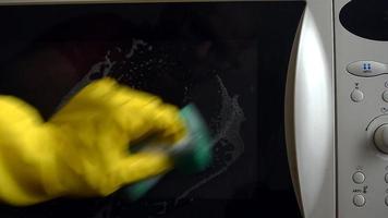 un hombre con guantes protectores lava la puerta cerrada del microondas con una esponja con una solución. limpieza húmeda en la cocina.