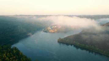 Zooming through fog to misty river, with vessels and touristic complex on bank. Aerial view of scenic valley with calm foggy river flowing between woodland hills in summer. Concept of landscape video