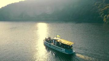 ferry pour passagers naviguant lentement sur la surface calme de la rivière le long des rives vallonnées par temps ensoleillé. vue aérienne du ferry avec drapeau bleu et jaune sur le dessus, flottant sur une rivière large et ondulée. concept de loisirs video