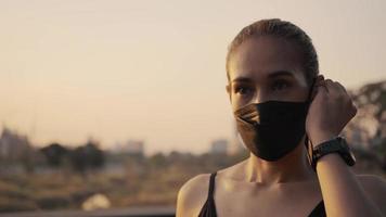 femme en vêtements de sport portant un masque de protection et mettant des écouteurs sans fil avant de commencer à faire du jogging dans la ville au coucher du soleil.