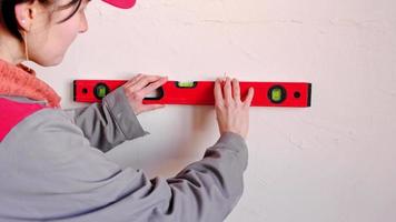 Drawing horizontal markings on the wall by level - DIY home repair. A woman in a red baseball cap and construction uniform is preparing the finishing of the walls video