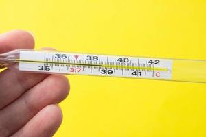 Mercury thermometer in hand on yellow background. Close up photo. Thermometer shows a temperature of 38.7 degrees Celsius photo