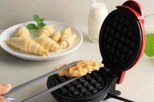 tome croissant wafle o croffle de la fabricación de wafles eléctricos con pinzas. horneado casero en la cocina, enfoque seleccionado. croffle popular en corea del sur foto