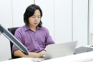 chico asiático de cabello largo se enfoca y se concentra en su trabajo frente a la computadora portátil en la oficina. foto