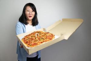 una joven asiática con anteojos abre una caja de pizza y se emociona con la pizza de gran tamaño que contiene. disparar con luz de estudio. foto