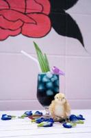 a yellow little chick with butterfly pea water and ice in a champagne glass is decorated by orchid and pandan leaf. It's shown and displayed on a pastel pink wood table in front of the cement wall. photo