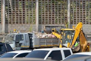 Yellow digger car is raising up polluted waste into garbage truck at parking lot with cement wall behind. photo