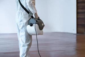 Professional technical man in prevention suit sprays sterilising solution by electrical spray machine on wood floor and white background with studio light. photo