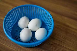 4 white chicken eggs in blue plastic basket on wood floor. 1 of 4 Chicken is ready to break out the egg shell from inside. photo