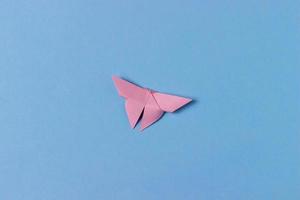 Pink origami butterfly folds out of paper. In center of a blue background. Education, pastime, hobbies, activities with children. Minimalistic background photo