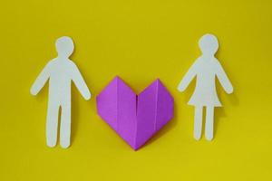 Two silhouettes of man and woman are carved from white paper with purple origami heart between them on a yellow background. The concept of love, relationships, family photo