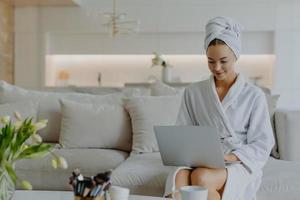 Horizontal shot of pretty young woman dressed in bathrobe and towel after shower sits on comfortabe sofa in living room works on laptop computer has own beauty blog types feedback to followers