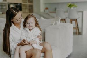 Beautiful brunette affectionate mother holds small curly haired cute dauhter in dressing gown pose in cozy room against modern apartment interior. Mom and little girl at home after taking shower photo