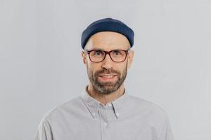 Headshot of handsome unshaven man wears transparent glasses, headgear, formal shirt, looks directly at camera, isolated over white background. Caucasian male with stubble dressed in fashionable outfit photo