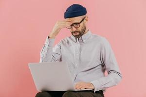 Male entrepreneur tries to make solution, keeps hand on head, tries to prepare financial report, focused in laptop computer, poses against pink background. Unshaven young man browses website photo
