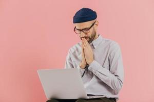 Professional male programmer keeps hans in praying gesture, looks at screen of laptop computer, believes in successful result of his work, wears formal shirt and hat, isolated over pink background photo