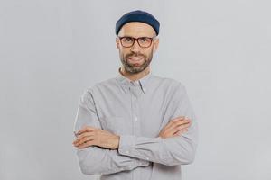 Horizontal shot of satisfied self confident male designer wears stylish headgear, dressed in white shirt, keeps arms folded, has dark stubble, isolated over white background. People and style concept photo