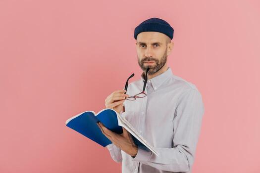 Self confident serious man with dark stubble, takes off spectacles, has attentive look at camera, holds opened textbook, learns necessary information, poses against pink background. Education photo