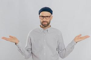 Photo of handsome man with uncertain expression, spreads hands, wears glasses and headgear, isolated over white backgrounnd. Unshaven young Caucasian male shruggs shoulders in bewilderment indoor
