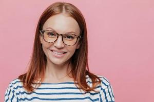 Close up shot of adorable young female student wears optical glasses, has tender smile, rejoices holidays wears striped clothes, models over pink background with copy space for your information. photo