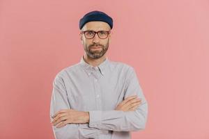 Self confident unshaven man crosses hands over chest, wears spectacles and white shirt, isolated over pink background. Bearded hipster ready for business meeting. People and fashion concept. photo