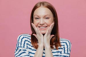 Photo of attractive European young woman has gentle smile, shows white teeth, touches cheeks with both palms, wears casual clothes, expresses sincere feelings, isolated over pink background.