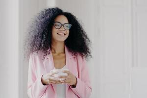 Photo of good looking African American prosperous busineswoman waits for partner in office, drinks coffee discusses future plans with colleague looks away with cheerful expression wears formal clothes