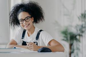 tiempo para estudiar. feliz adolescente de piel oscura sostiene un bolígrafo, escribe la información necesaria para el trabajo del curso, ocupada preparándose para las clases, vestida con ropa informal y espectáculos, trabajando duro foto