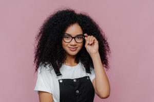 Pretty African American woman keeps hand on frame of glasses, smiles happily, wears white casual t shirt and black overalls, enjoys spare time, poses over violet background. Ethnicity, fashion concept photo