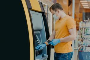 Man wears protective mask and medical gloves, uses ATM machine, prevents from virus pandemic, introduces pin code. Safe money withdrawal during coronavirus outbreak. Payment, influenza concept photo