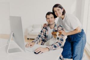 Happy husband and wife pose near computer, keyboard and surf information, work in domestic atmosphere with dog, pose in coworking space, connected to wireless internet. Lovely couple at desktop photo