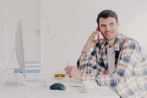 Horizontal shot of busy male freelancer has telephone conversation, dressed in checkered shirt, writes down information, sits at desktop with computer, solves financial problem, poses at workplace photo