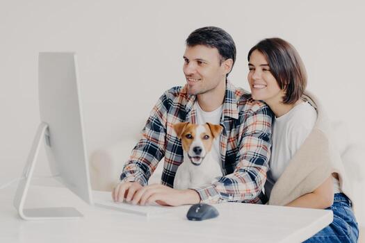 Happy friendly couple sit together with dog, make booking of hotel for future trip, keyboard and look at computer, chat online, pose at white desktop, connected to free wifi, browse internet together photo