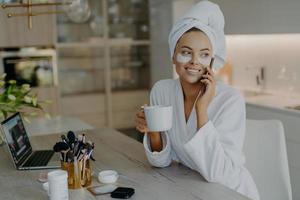 Pretty smiling woman applies hydrogel patches under eyes enjoys grooming herself after shower dressed in bathrobe drinks tea has telephone conversation sits in chair at home. Face care procedures photo