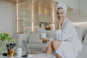 Cheerful refreshed woman with healthy skin wears bathrobe after taking shower drinks coffee and looks thoughtfully aside sits on comfortable sofa works on laptop computer uses cosmetic products photo