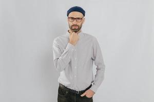 Waist up shot of serious man keeps one hand under chin, other in pocket, has stubble, wears hat, white shirt, trousers, isolated over white background, listens something attentively. Masculinity photo