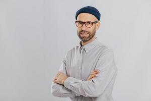 Isolated shot of pleasant looking unshaven male keeps hands crossed, wears stylish hat, optical glasses, models over white background with copy space for your advertising content, enjoys leisure photo